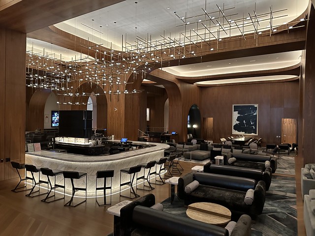 Signia by Hilton San Jose lobby with modern light installation of crystal glass and metal rods suspended from ceiling. A large white marble bar with bar stools is a focal point in the first floor lobby. A bank of dark leather sofas with black and white accent bolsters are placed throughout the cavernous room with dark wood paneled walls. 