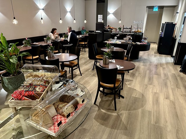 Best Western Dam Square breakfast room with 8 circular wood tables. Two hotel guests sit at a table, while a third gentleman sits alone, to the right of their table. Bread and crackers are on display upon entrance to the room. A buffet counter is toward the back of the breakfast room. 