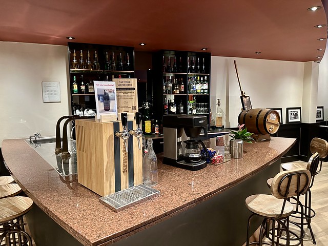 Hotel bar with liquor bottles, water tap dispenses still and sparkling water. To the right is a coffee maker and a small barrel of wine on the counter. Three wooden bar stools are under the counter. 