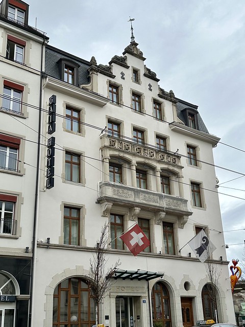 White exterior of 5-story GAIA Hotel Basel. The red flag of Switzerland and white flag hang above the entrance to the Swiss hotel. 