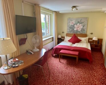 Double bed with white sheets and red duvet with red velvet bench at end of bed. Busy pattern of gold on red carpet with wood desk to the left of hotel room and one red desk chair. Above the bed is a large oil painting of a flower. A flat screen tv is above the desk, mounted on the wall. A small, white portable fan is on the right of the desk.