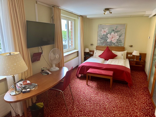 Double bed with white sheets and red duvet with red velvet bench at end of bed. Busy pattern of gold on red carpet with wood desk to the left of hotel room and one red desk chair. Above the bed is a large oil painting of a flower. A flat screen tv is above the desk, mounted on the wall. A small, white portable fan is on the right of the desk. 