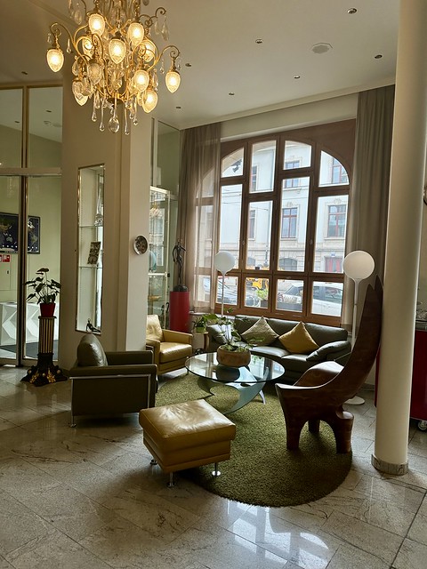 Olive green leather sofa against large picture window looking out to Basel railway station. Mustard yellow leather and olive green leather chairs surround circular glass table top with modern stainless steel , freeform base of coffee table. A small potted plant is on glass tabletop. A green oval area rug is under table with marble flooring. Above the table is an antique glass chandelier. 