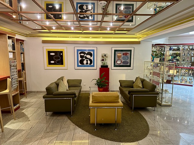 Sitting area in GAIA Hotel Basel with 2 olive green leather sofas and mustard yellow leather chair around a square glass table. A single green plant in a red planter is on the table. Across the back wall are three framed modern art pieces. The reflection of the artworks shines above, on the glass ceiling. 