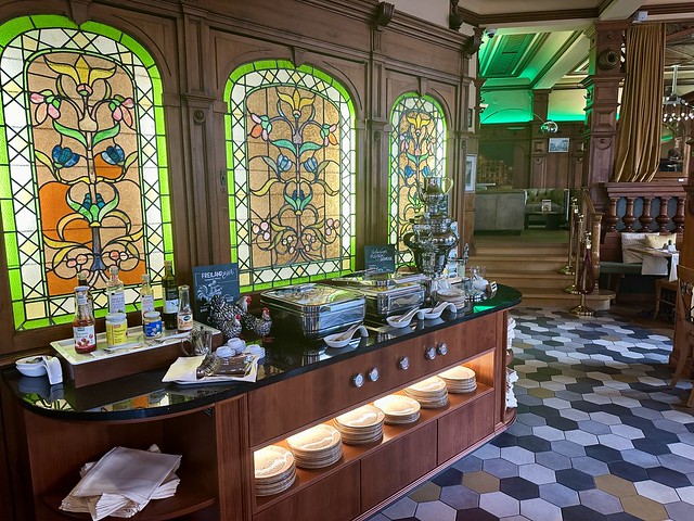 Three antique leaded, stained glass windows are above the breakfast buffet station. Two stainless steel chaffing dishes hold eggs and meats. A large, silver dispenser holds hot water for tea and dishes are stacked below in the wood buffet cabinet. 