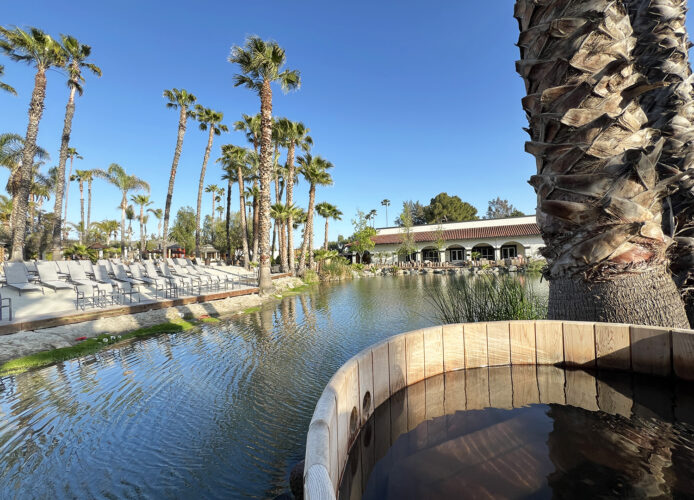 Round wooden soaking tub next to a lake with sandy beach and lounge chairs and palm trees