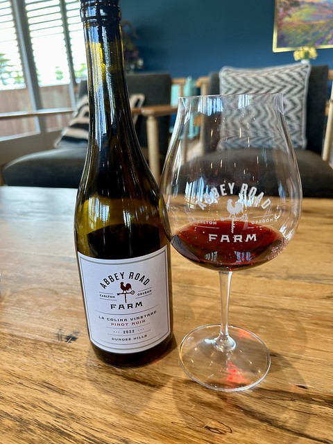 Abbey Road Farm bottle of La Colina Pinot Noir and logo Pinot Noir red wine glass with Abbey Road Farm La Colina Pinot Noir poured in the glass. Bottle and glass on a oak plank table in the winery tasting room. 