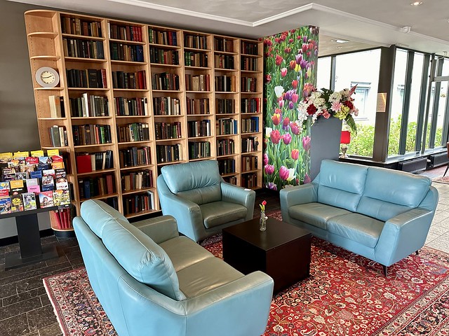 Floor-to-ceiling wooden bookshelves line a section of the hotel lobby to create a reading area. Two light-blue leather sofas, as well as a light blue leather chair surround a square black table. A lone red tulip is on the table in a vase. 