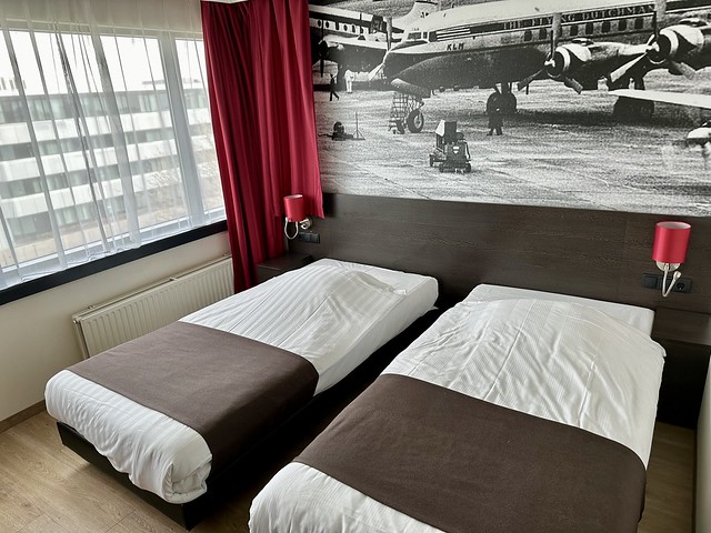 Amsterdam Airport Hotel room with 2 single beds. One pillow is tucked under the white sheet on each bed. A small red lamp with electrical plug in is on either side of the beds. 