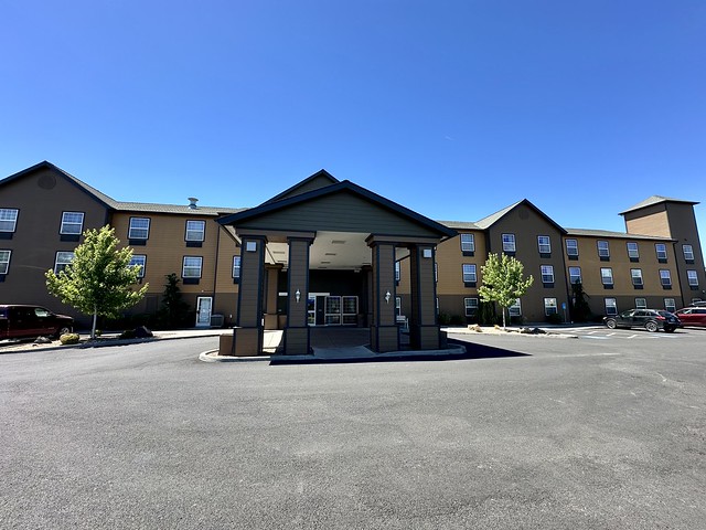 Front exterior and drive through portico of the Best Western Plus Bend North 3-story hotel. Brown colored exterior with a single tree on either side of hotel entrance. 