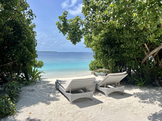 Two wicker beach chairs on white sand in front of aqua water.