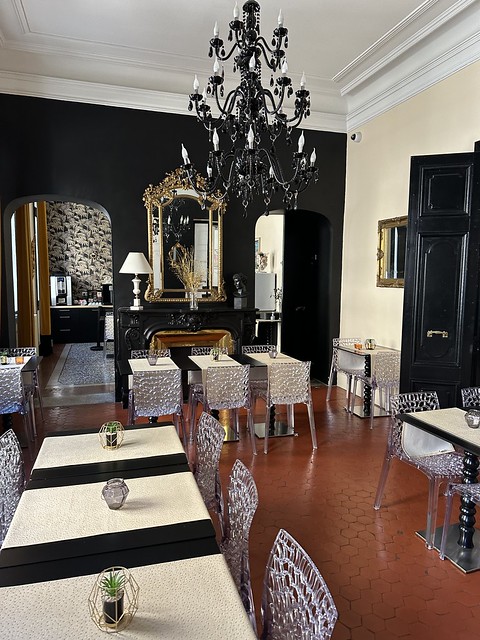 Hotel La Residence breakfast room with 8 small tables with plastic chairs, each seating 2 persons. A black chandelier hangs from the ceiling. 