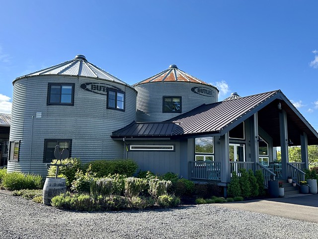 Two former Butler grain silos are welded together (3rd silo not shown) to create Silo Suites Bed and Breakfast in Oregon. Entrance to B&B has dark metal roof with three wood steps, leading to lobby. Gravel road and greenery in front of building.