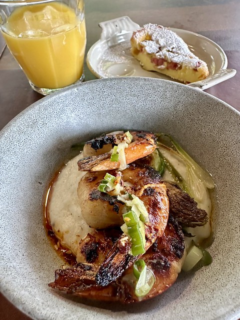 Ceramic bowl holding three glazed shrimp, resting on a bed of grits. Left, upper corner of bowl is glass of orange juice. Right of bowl is a slice of French pastry dusted with powdered sugar on a separate small white plate. 