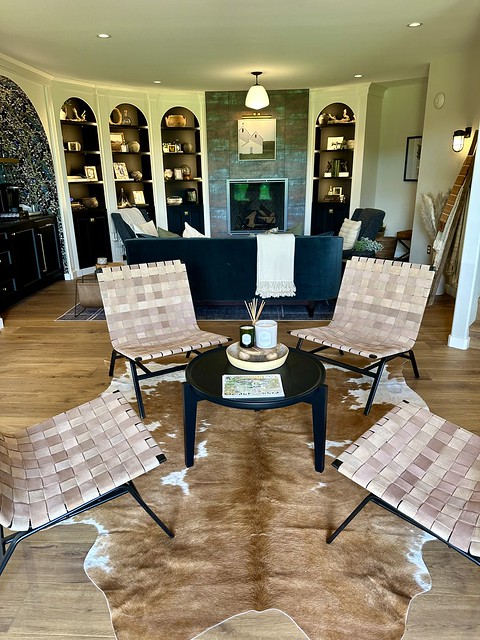 Silo Suites lobby with 4 black steel and beige woven leather chairs surround a small black circular table. A brown and white cowhide rug is under the table and chairs. Behind the chairs is a blue velvet sofa and 2 blue velvet chairs on either side of the couch. A gas fireplace and wall are beyond the sofa. Either side of the fireplace is shelving with 3 decorative selves, left of fireplace and one built-in shelf to the right of fireplace. Shelves are filled with decorative books, plants and art. 