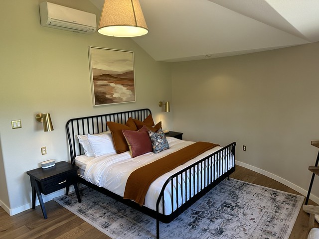 King bed with white duvet, black iron bedframe. Five brown leather and cloth print decorative pillows on bed. brown blanket as runner at base of bed. Hardwood plank floor with geometric patterned area rug under king bed. Abstract painting above bed and 2 wall mounted brass reading lamps mounted at either side of king bed. 