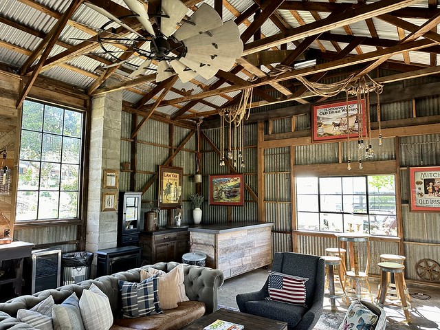 The Tool Shed at Silo Suites is a corrugated metal barn room, filled with a brown leather sofa and grey fabric chair next to sofa. Partial wood table showing, with magazine on top. Right of sofa is high top circular bar table with 4 bar stools surrounding table. Back right corner is wood table with metal top that serves as bar area for entertaining. Old Irish Whiskey art advertisements are framed on the walls to give room a bar feel. 