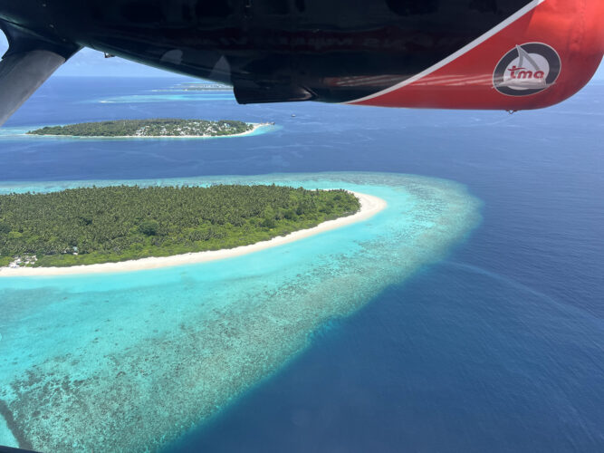 View out the window of a seaplane of lush island surrounded by white sand and aqua water