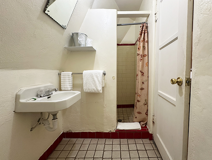 A small crowded bathroom with worn tiles features a small wall-mount sink and tiny walk-in shower