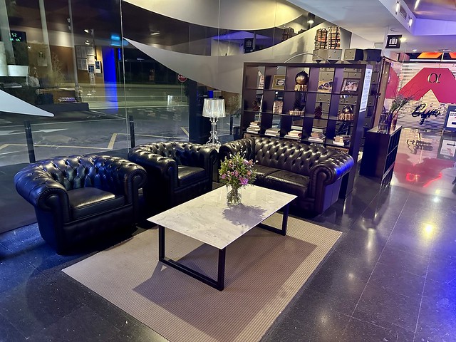 Hotel Alfa Aeropuerto lobby with 1 brown leather sofa and two brown leather chairs next to a rectangular white granite coffee table. Coffee table has small bouquet of fresh, spring flowers in clear vase. 