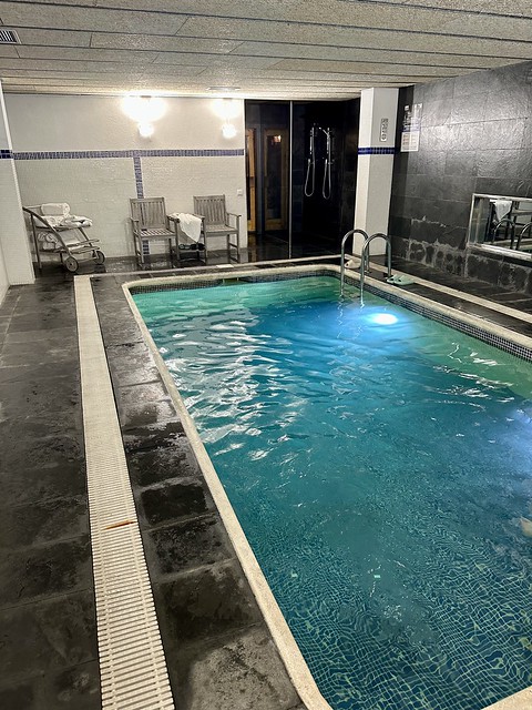 Indoor, rectangular-shaped hotel swimming pool. Two chairs with a towel cart are at end of pool. A shower stall is by pool for guests to use before entering the pool. 