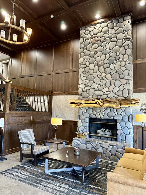 Floor to ceiling river rock stone fireplace inside the hotel lobby. One chair and camel-colored 2-person sofa are across from the fireplace. A large, square dark wood table sits upon a dark area rug in the lobby in front of fireplace. Wooden stairway leads to 2nd floor of the hotel. 