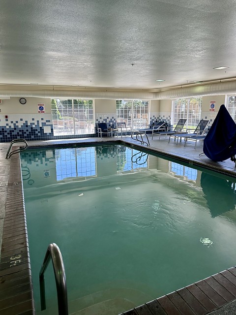 Indoor hotel swimming pool with metal handrail. An ADA compliant chair was covered with a blue tarp beside the pool. 