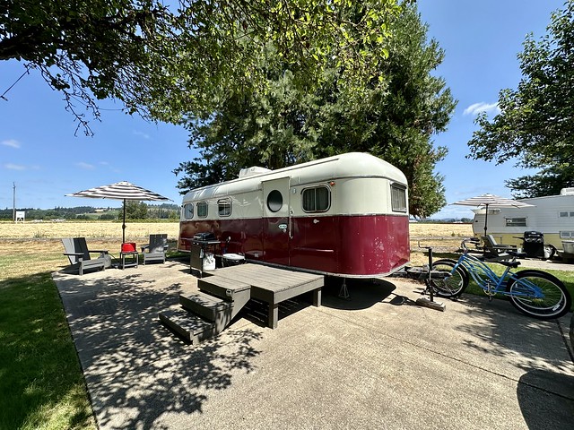 1951 M System vintage trailer with cream and red coloring. The trailer has 2 Adirondack chairs and a patio table & cloth umbrella. Two blue cruiser bikes are to the right of vintage trailer. 