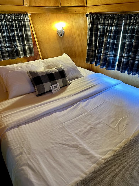 Queen bed inside 1951 M System vintage trailer. White bedding, 2 white pillows and a decorative rectangular black and white pillow are on top of the queen bed at Vintages Trailer Resort.