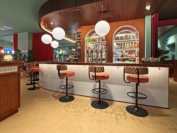 A curved bar with a glass tile front, earth-tone bar stools, hanging globes and a wall of drink bottles