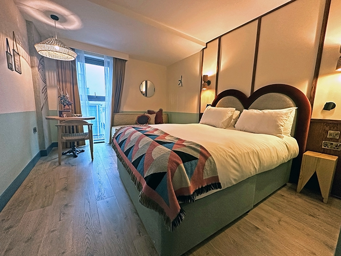 A guest room at Room2 Belfast with a pale green, burgundy and white color palette. A large bed is in the middle of the room and small round table and chair is by the window.