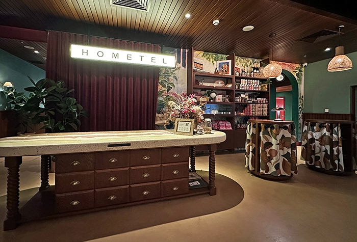 An illuminated sign says Hometel hangs above a chunky wood check-in counter at Room2 Belfast. To the side is a small market wall.