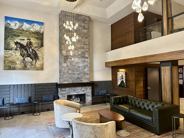 The 2-story Newberry Hotel lobby features a large painting of a cowboy on a black horse with the Central Oregon mountain peaks behind him. To the right, a stone 2-story fireplace rises to the ceiling against a white wall. Two beige chairs are across from a small, wood oval coffee table and a black leather sofa. Two modern glass tear-drop chandeliers suspend from the hotel ceiling into the lobby. 