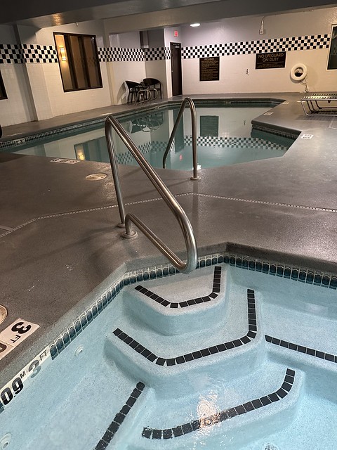 Indoor hot tub and separate swimming pool. Three stairs lead to the hot tub with a metal handrail. Beyond is the pool with a metal handrail. 