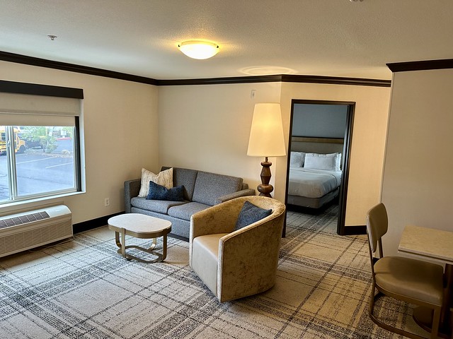 Newberry Hotel king suite with grey cloth sofa bed, small oval table with granite countertop. An oval beige fabric and leather swivel chair is next to the sofa. A rectangular window looks out to the parking lot. Beyond the chair is the separate king bedroom. Partially shown is a small table and chair in the hotel suite. 