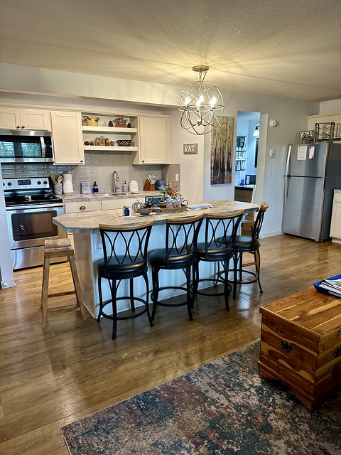 The kitchen includes a full size stainless steel refrigerator, oven with microwave oven above stovetop. Therre is a sink and dishwasher. The kitchen counter has 4 chairs and 2 wood stools at either end of the island counter. 