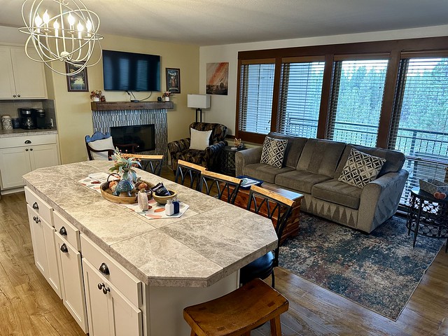 Kitchen with white quartz countertop. Gray cloth sofa with 2 throw pillows is adjacent to kitchen counter. Four full-length glass picture windows lead out to the balcony, overlooking the pine forest. A brown leather chair and floor lamp are to the left of the sofa and right of the electric fireplace. A flatscreen tv is above the fireplace.