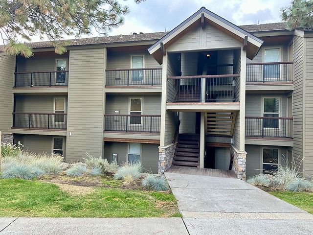 Three-story building with six hotel rooms per building. Walk upstairs to second and third floor. Rectangular large window on each of the three floors. Cement walkway and small patch of green grass and natural grass to the left and right of cement walkway. 