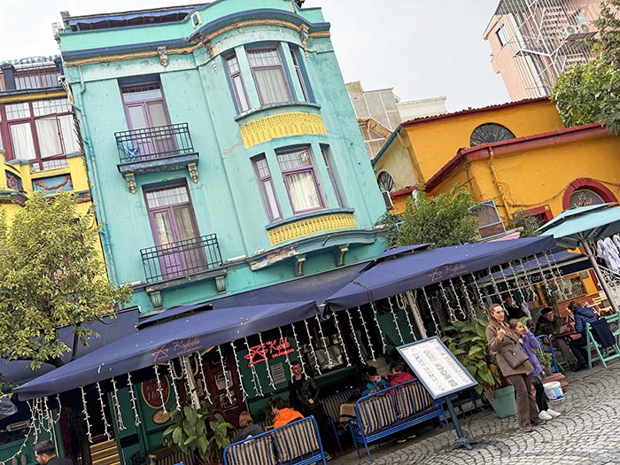 Exterior of narrow townhouse-style turquoise building with restaurant seating in front on sidewalk