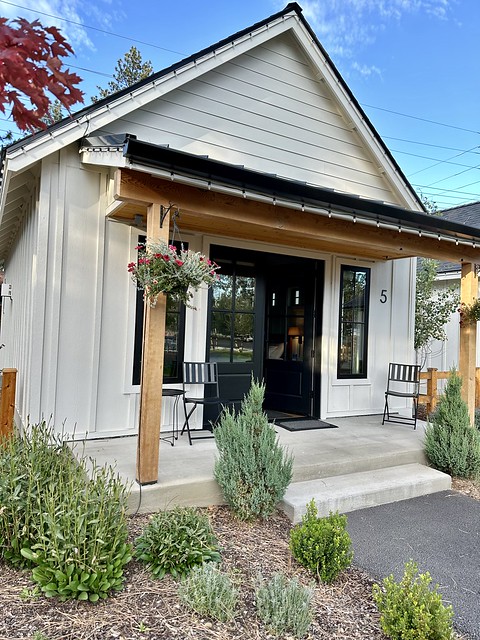 The Scout Cottage #5 exterior, painted cream with black trim and 2 wood posts on the front porch. The concrete front patio has a black chair on either side and a small round patio table. A seasonal hanging plant basket is on the left of the patio. Some small drought-tolerant plants are in the ground in front of cottage. 