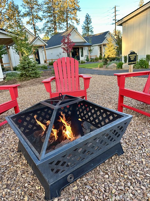 The outdoor fire pit is surrounded by three bright bred Adirondack chairs. Behind the fire pit, with small rocks are the Scout Cottages. 