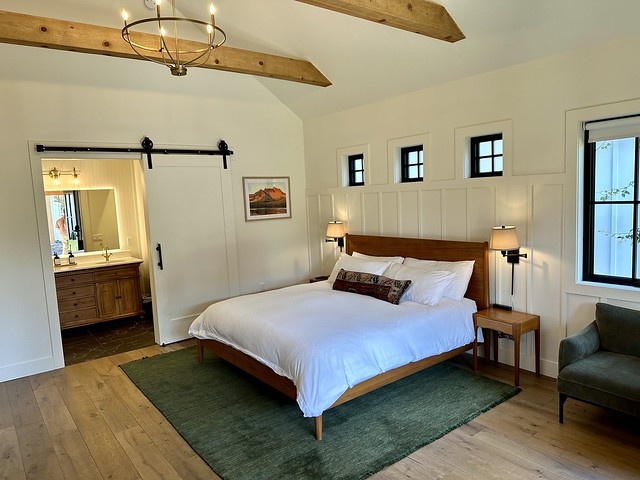 King bed with white duvet and area rug under bed. Three small square windows are above the bed. A sliding barn door opens to the guest bathroom. The walls are painted white with exposed wood beams in the ceiling. 