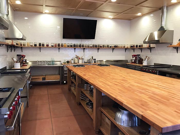 A state of the art demonstration kitchen with stainless appliances, a massive center island, shelves lined with jars of herbs and spices, and a TV on the wall