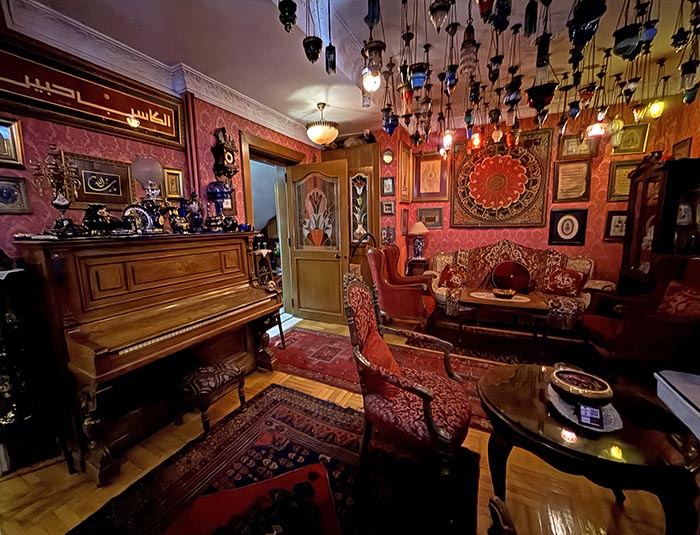 Red sitting room filled with antique furnishings, piano and dozens of glass hanging ceiling lamps